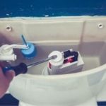 Hand holding screwdriver adjusting toilet tank components, including flapper and fill valve, relevant to preventative maintenance for running toilets.