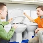 Two men discussing toilet components and maintenance, with one demonstrating the inner workings of a toilet tank, relevant to plumbing services and preventative maintenance tips.