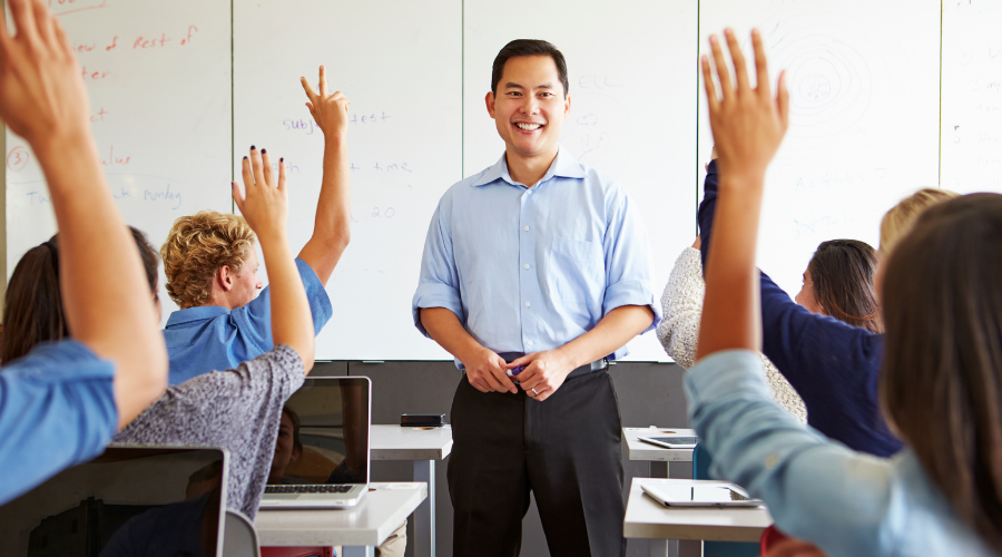 Teacher engaging with students in a classroom, students raising hands to participate in discussion.