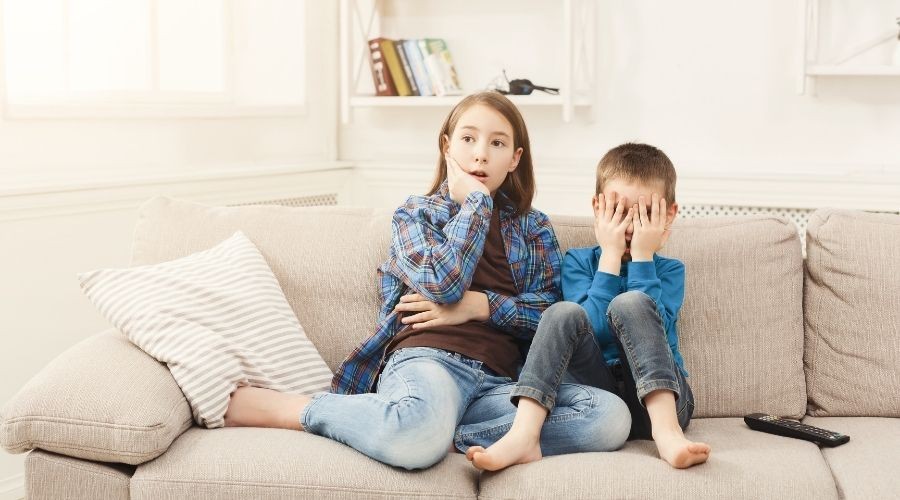 Children watching a scary movie on the couch, with one girl looking surprised and a boy covering his eyes, illustrating tips for a spooky movie marathon.