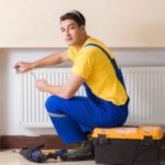 HVAC technician in blue overalls and yellow shirt servicing a radiator, with tools and a toolbox nearby, representing reliable heating maintenance services.