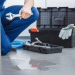 Plumber kneeling on wet floor with wrench, surrounded by tools, indicating urgent leak repair needs.