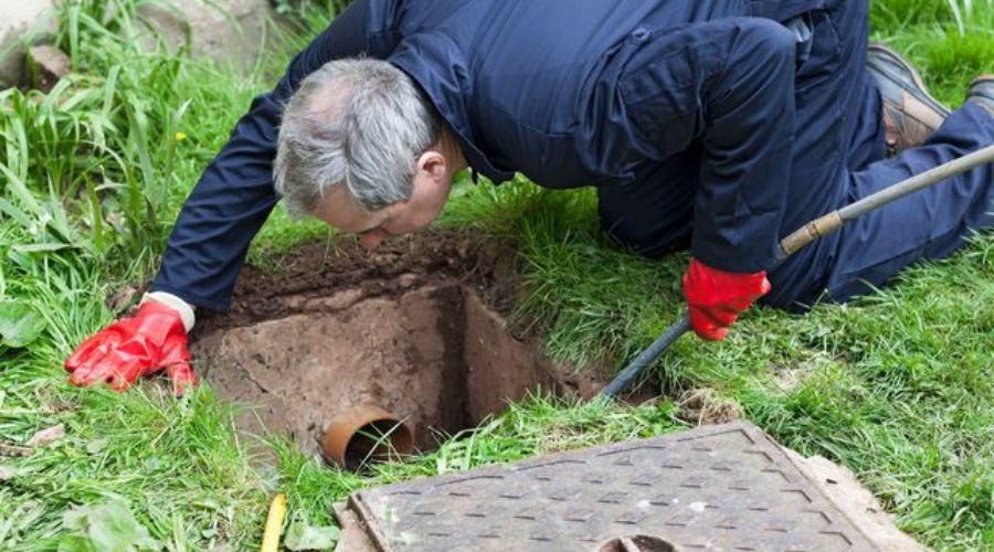 Plumber inspecting outdoor plumbing system, digging near a sewer line in grassy area.